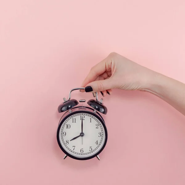 Creative flat lay morning time emotions concept top view of woman hand holding black vintage alarm clock pastel millennial pink color paper background with copy space minimal style square template