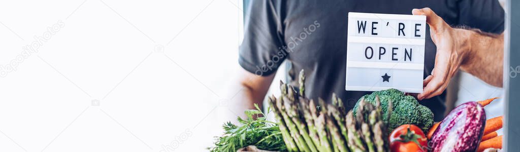 End of quarantine lockdown. Man holding lightbox with greeting text message We're open and Fresh greens and vegetables box. Welcoming grocery shop clients after coronavirus Covid-19 pandemic outbreak