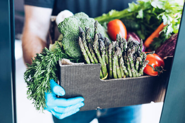 Fresh organic greens and vegetables delivery during coronavirus Covid-19 pandemic outbreak. Man hands wearing blue latex medical gloves holding box with safe grocery delivering in the house doorway
