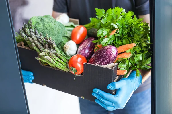 Fresh organic greens and vegetables delivery during coronavirus Covid-19 pandemic outbreak. Man hands wearing blue latex medical gloves holding box with safe grocery delivering in the house doorway