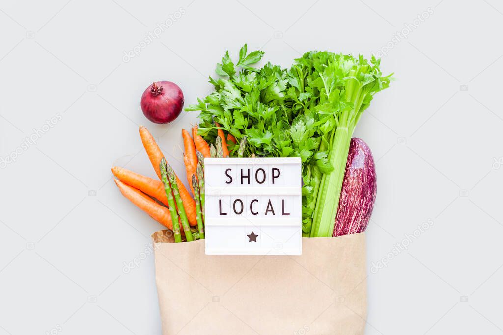 Fresh organic vegetables in eco craft paper shopping bag with text Shop Local on lightbox flat lay, top view with copy space on gray background. Sustainable lifestyle. Zero waste, plastic free concept.