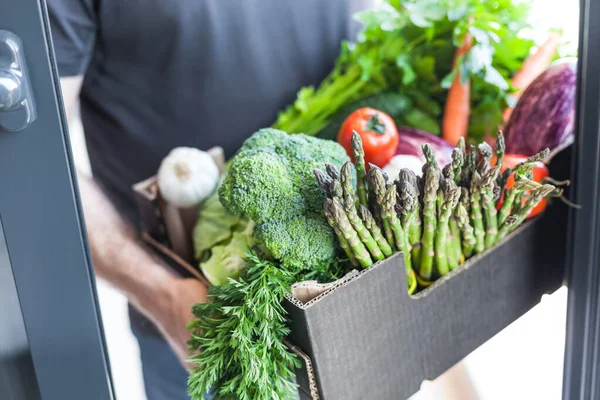 Fresh organic greens and vegetables delivery. Man hands holding box with farmer bio crop delivering in the house doorway. Small local business support. Online grocery shopping