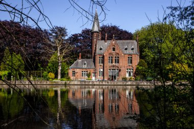 Minnewater gölü panoraması, gotik yapının yansıması ve Castle de la Faille yakınlarındaki kafede insanlar, bulutlu mavi gökyüzü, Bruges, Belçika