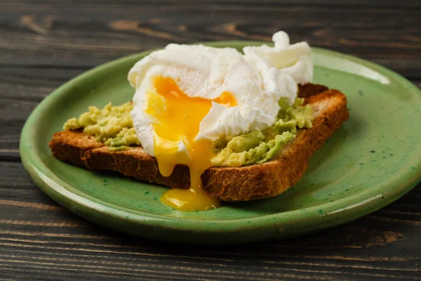 Wholemeal bread toast and poached egg with avocado on green plate on wooden table
