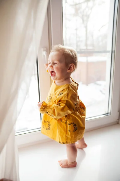 Niña pequeña de 9 meses de pie en el alféizar de la ventana  . — Foto de Stock