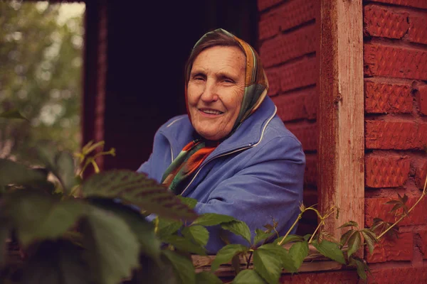 old grandmother in   handkerchief sitting on   porch of   wooden house.