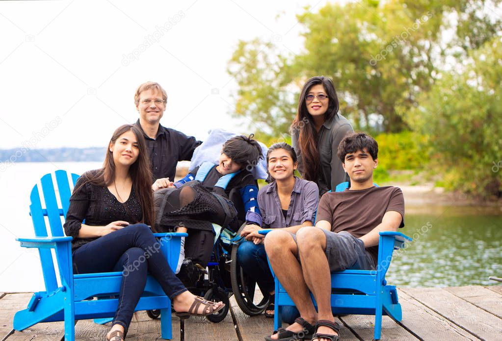 Multiracial family with special needs child sitting outdoors together on summer day. Child is sitting in wheelchair.