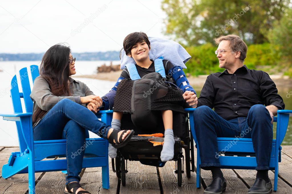 Multiracial couple sitting with disabled twelve year old son in wheelchair while sitting in blue adirondack chairs on wooden pier by lake on summer day