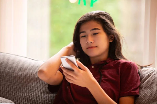 Biracial adolescente menina sentada no sofá cinza olhando para smartphone — Fotografia de Stock