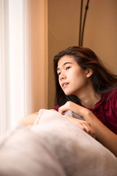 Biracial teen girl sitting on gray couch looking out window — Stock Photo, Image