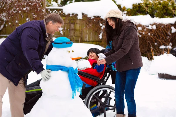 Menino deficiente na cadeira de rodas construção boneco de neve com a família durante — Fotografia de Stock