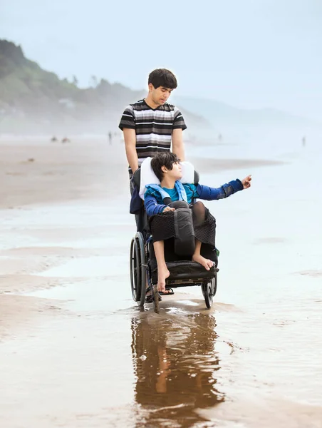 Молодой человек толкает брата-инвалида в инвалидной коляске на пляже — стоковое фото