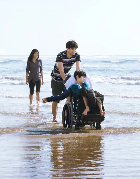 Rodzeństwo spaceru niepełnosprawny brat na wózku inwalidzkim w wodzie wzdłuż b — Zdjęcie stockowe