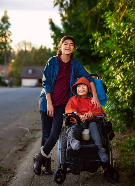 Sorella in piedi accanto a disabile fratellino in sedia a rotelle ou — Foto Stock