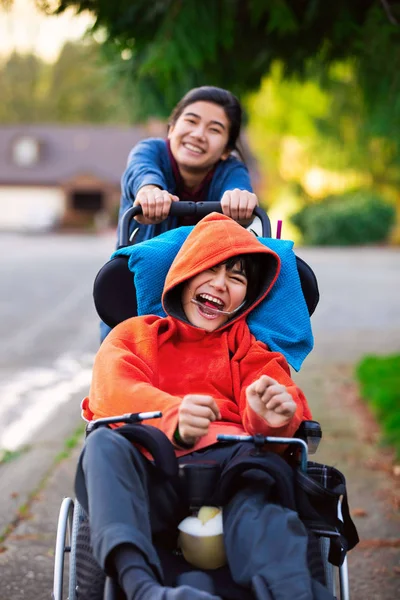 Sister pushing disabled little brother in wheelchair around neig — Stock Photo, Image