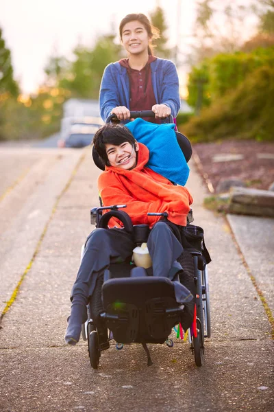 近所のまわりで車椅子で無効にされる弟を押している姉妹 — ストック写真