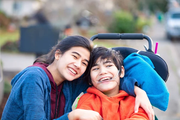 Старшая сестра обнимает брата-инвалида в инвалидной коляске на улице, улыбается — стоковое фото