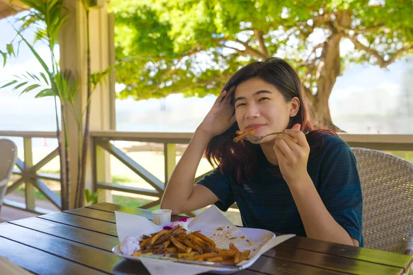ホノルルのワイキキのビーチ沿いの屋外レストランに座っているアジア系の10代の女の子の笑顔晴れた日の午後にフランスのフライドポテトを食べる — ストック写真