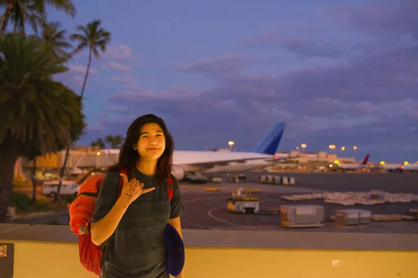ハワイ空港に立つ10代の少女は 夕方のフライトの前にシャカのサインをしています 背景に暗い空と飛行機 — ストック写真
