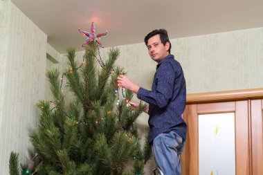 Yakışıklı orta yaşlı adam bir yıldız ve salon iç merdiven üzerinde duran renkli ampuller çelenk ile Noel ağacı süsle. Yeni yıl tatil kavramı, rahat ev