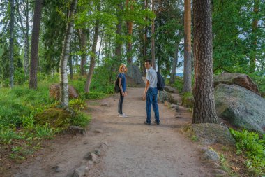 Orta yaşlı adam esmer ve Kızıl saçlı Genç Bayan orman yolu yürüyün. Güzel manzara arka plan üzerinde turistler. Monrepos Park, Vyborg, Rusya Federasyonu. Seyahat kavramı