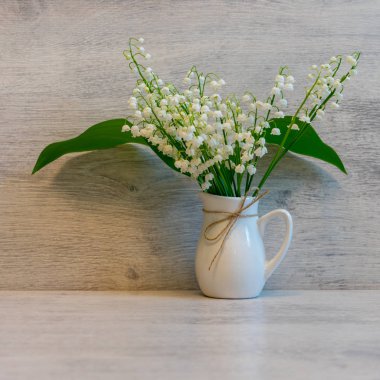 Hafif ahşap arka planda, küçük bir sürahi içinde, vadi çiçeklerinin taze zambakları. Güzel bahar natürmortu, tebrik kartı için hazırlan, boşluğu kopyala