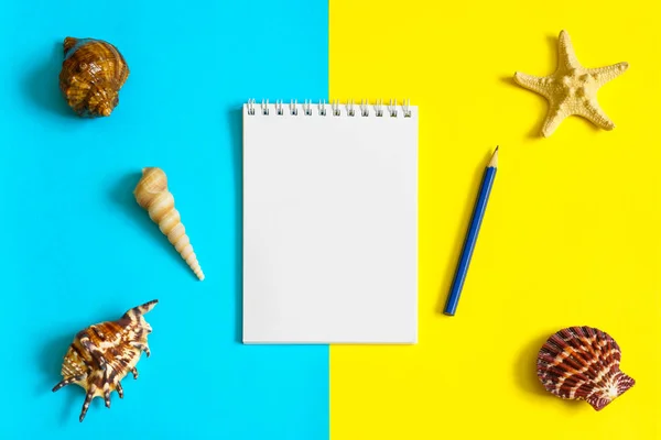 Notebook and pencil with seashells and sea stars on blue and yellow paper background. Mock up, flat lay, copy space, top view. Summer vacation concept