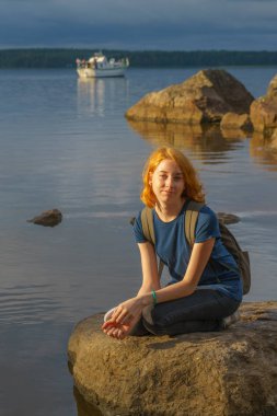Genç ve güzel kadın Kızıl saçlı oturan bay shore yakınındaki taş yaz akşamında. Güzel manzara arka plan üzerinde turist. Monrepos Park, Vyborg, Rusya Federasyonu. Seyahat kavramı