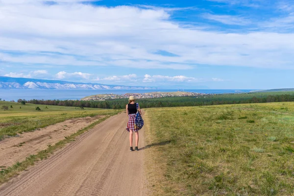 Interpoleringen turist flicka i hatt och ryggsäck promenader på land vägen ensam och beundrande pittoreska landskap av Bajkalsjön, byn och bergen, blå himmel. Resa, vandring och sommaren semester koncept — Stockfoto