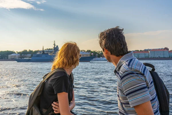 Middle-aged dark-haired man and young redhead lady on Neva river embankment looking at warships and architectural ensemble in summer evening at sunset. Travel concept. Saint Petersburg, Russia
