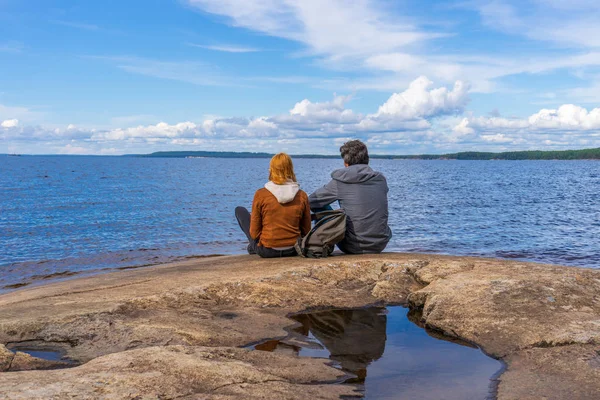 Tourists man and woman sitting on northern lake shore in summer day. People relaxing and admiring beautiful landscape. Travelling and discovering distant places of Earth. Onega lake, Karelia, Russia