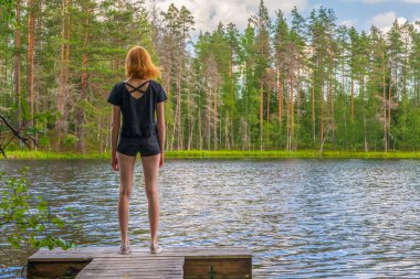 Kuzey göl ve ormanın güzel peyzaja karşı yaz gününde ahşap dalgalı mamülleri üzerinde duran sevimli zencefil kız. Dünya 'nın uzak yerlerini keşfetmek ve seyahat etmek. Karelia, Rusya Federasyonu