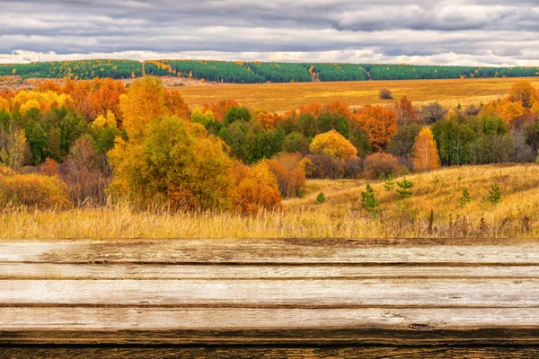 Empty wooden table with blurred picturesque autumn landscape of panoramic view from hill to lowland with field and grove in cloudy day. Mock up for display or montage products