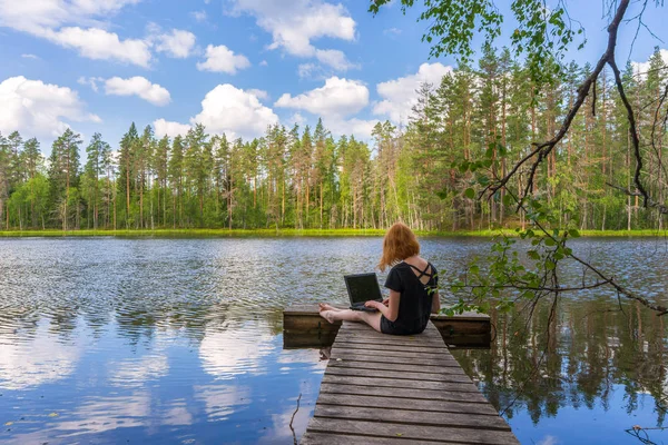 Roztomilá zázvorná dívka, která sedí na dřevěné lávce a pracuje s laptopem v letních dnech proti krásné krajině severního jezera a lesa. Koncepce na volné noze, práce a cestování. Karkaa, Rusko — Stock fotografie