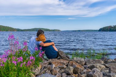Turistler in anaç erkek ve kızıl saçlı genç kadın güneşli yaz akşamı göl koyu kıyısında taşlar üzerinde oturuyor. İnsanlar ladoga gölünün güzel manzara dinlenme ve hayranlık. Karelia, Rusya Federasyonu