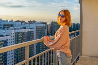 Pullover, kot pantolon ve güneş gözlüğü gün batımında yüksek katlı çok katlı konut binasına karşı balkonda duran tween redhead kız. Güzel bir görünüm, şık şehir sokak kıyafeti, genç moda