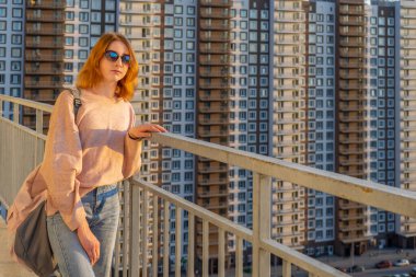 Pullover, kot pantolon ve güneş gözlüğü gün batımında yüksek katlı çok katlı konut binasına karşı balkonda duran tween redhead kız. Güzel bir görünüm, şık şehir sokak kıyafeti, genç moda