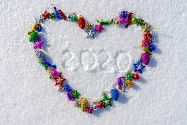 Cuore simbolo fatto di molti colorati Natale o giocattoli di Capodanno e il testo 2020 sulla neve fresca bianca nella soleggiata giornata invernale. Buon Natale e felice anno nuovo. Concetto vacanze invernali. Vista dall'alto, copia spazio — Foto Stock