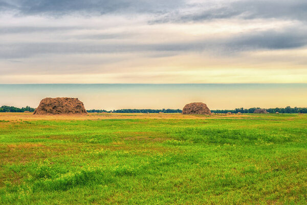 Picturesque summer landscape with beveled meadow and haystacks in cloudy morning. Hay harvest at farmland. Beautiful agricultural background.