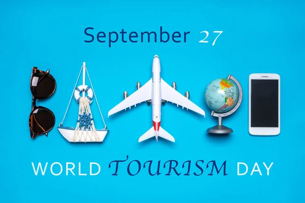 Happy world tourism day. Touristic objects, smart phone, sunglasses, globe and decorative airplane and vessel on blue background. Flat lay, top view. Text September 27, WORLD TOURISM DAY.