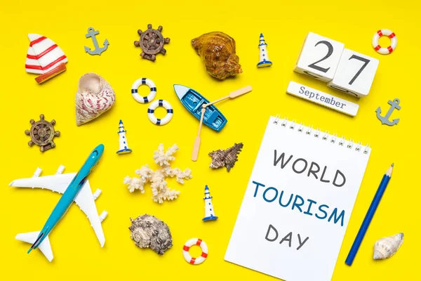 Happy world tourism day. Touristic decorative items, plane, vessel, seashells on bright yellow background. Flat lay, top view. Calendar date September 27, notebook with text WORLD TOURISM DAY.