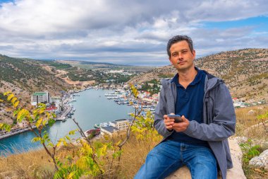Yakışıklı orta yaşlı turist bulutlu bir günde taşın üzerinde oturmuş küçük bir sahil kasabasının güzel manzarasına karşı. Tepelerin, denizin, gökyüzünün ve kasabanın arka planında akıllı telefon kullanan sıradan giysili bir adam..