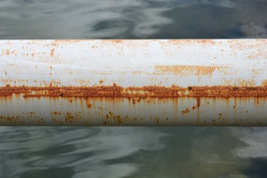 Belirgin yatay pas çizgisi olan paslı beyaz metal bir borunun yatay yakın çekim görüntüsü bulanık bir su kütlesi üzerine kuruludur..