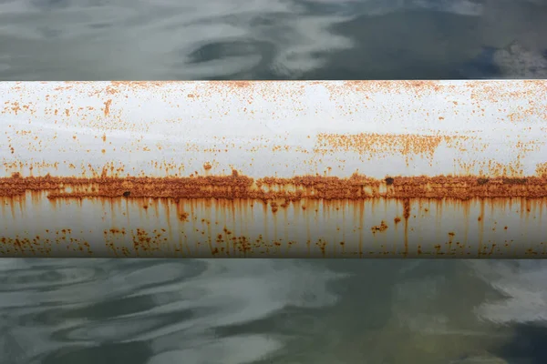 Belirgin yatay pas çizgisi olan paslı beyaz metal bir borunun yatay yakın çekim görüntüsü bulanık bir su kütlesi üzerine kuruludur..