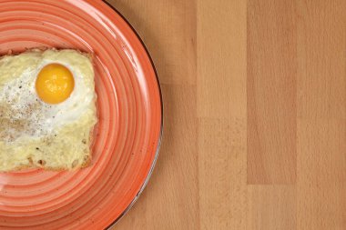 Tahta bir yüzeyin sol tarafına yerleştirilmiş turuncu bir tabakta peynirli ve kızarmış yumurtalı tost.