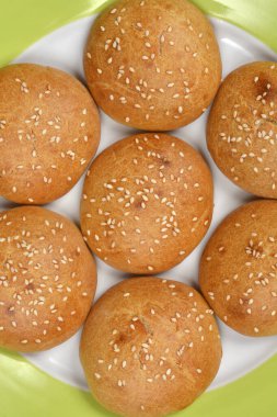 Yakın plan, tepeden tırnağa yedi taze susamlı çöreğin parlak yeşil kenarlı beyaz bir tabağa dizilmiş hali.