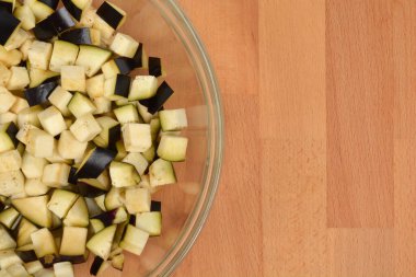 A top-down close-up view of freshly diced eggplant pieces filling the left side of a clear glass bowl on a wooden table with copy space