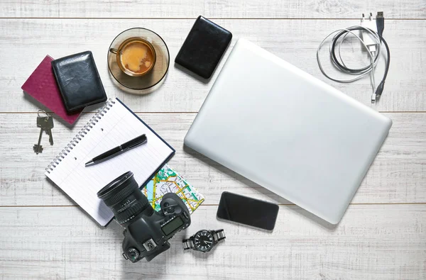 View of a wooden table with a laptop, external hdd, usb cable, charger, mobile, notebook, camera, pen, watch, wallet, passport, map, keys and a cup of coffee on it