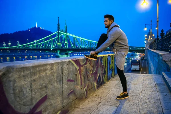 A handsome young sportsman ties his shoelace on the street at night on the riverside with a bridge behind him