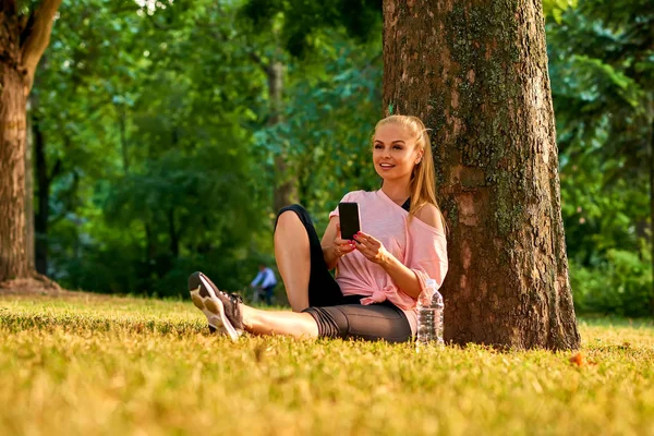 Hermosa Joven Deportista Sentada Árbol Parque Atardecer Mientras Usa Teléfono —  Fotos de Stock